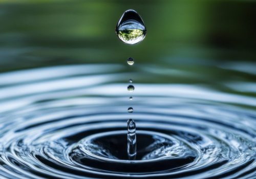 A drop of water falling into a large body of water. The water is calm and still, with no ripples or waves. The drop of water is the only thing moving in the scene, creating a sense of stillness